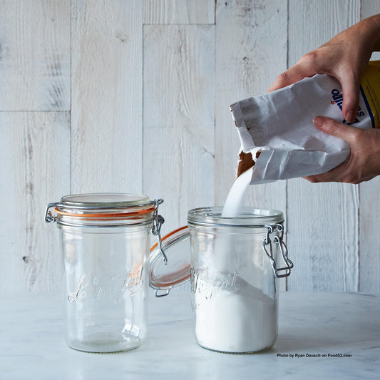 Two glass Le Parfait jars being filled with white sugar against wood background